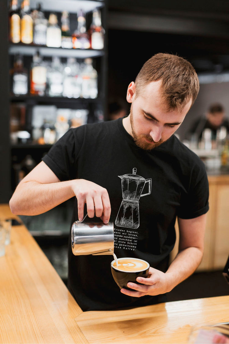 „Barista mit schwarzem Shirt gießt Milch in Kaffee – Latte Art entsteht. Shirt zeigt Mokkakanne und Zitat über Frieden durch Zuhören und Kaffee.“