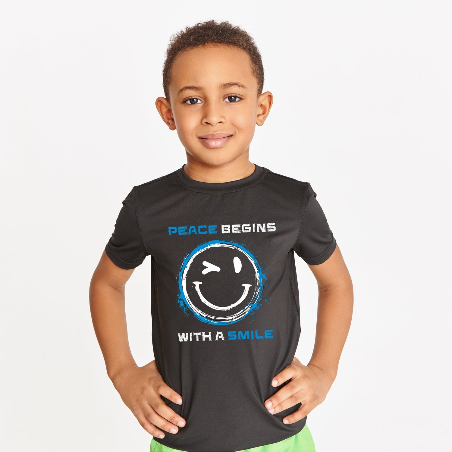 Dunkelhäutiger Junge in schwarzem T-Shirt mit dem weißen Schriftzug Peace begins with a smilie und einem augenzwinkernden Smiley – steht mit selbstbewusster Haltung, die Hände in die Hüften gestemmt.“