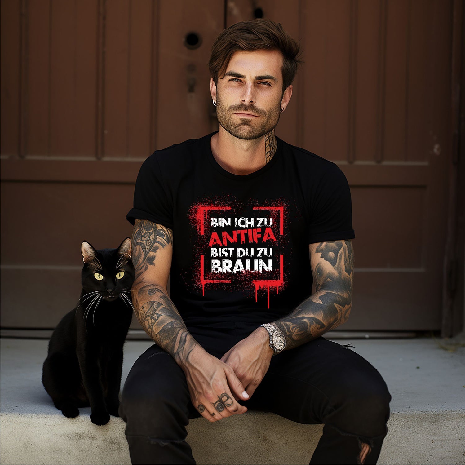 Mann mit Tattoos sitzt neben einer schwarzen Katze auf Treppenstufen, trägt ein Shirt mit „BIN ICH ZU ANTIFA BIST DU ZU BRAUN“.